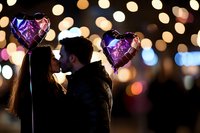 Romantic silhouette of couple kissing with heart-shaped purple balloons against blurred bokeh lights background, creating magical Valentine's Day atmosphere.