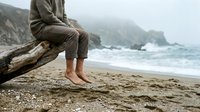 Person sitting on driftwood log at misty coastal beach with waves, barefoot relaxation and contemplation by ocean shore.
