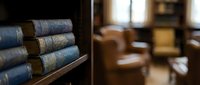 Antique leather-bound books stacked on wooden shelf in classic library setting with warm lighting and blurred interior background.