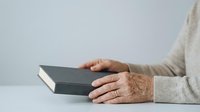 Senior woman with age spots and wrinkles wearing beige sweater holding dark hardcover book on white surface and background.