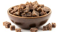 Dark brown sugar cubes in ceramic bowl with scattered pieces on white surface, natural sweetener close up view showing crystalline texture and rich caramel color.