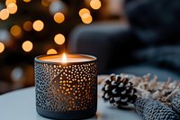 Glowing decorative candle holder with intricate pattern casting warm light and shadows, pine cones on table against blurred bokeh background for holiday decor.