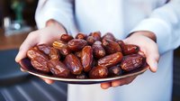 Fresh organic medjool dates served on dark ceramic plate by person in white clothing, healthy natural dried fruit snack for Middle Eastern cuisine.