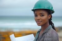 Young African American female construction engineer in green hard hat reviewing blueprints at coastal construction site, displaying confidence and expertise in professional setting.