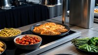 Professional kitchen setup with grilled chicken breast, pasta, cherry tomatoes and fresh vegetables on modern stainless steel counter for culinary preparation.
