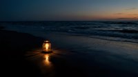 Glowing lantern on wet sand beach at twilight with ocean waves and dramatic sunset sky creating peaceful evening atmosphere for relaxation and meditation.