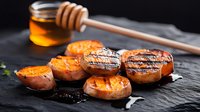 Grilled sweet potatoes with honey glaze on black slate, showcasing caramelized edges and golden-brown grill marks against dark background.