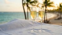 Two champagne flutes filled with sparkling wine on white table at tropical beach sunset with palm trees and ocean view for romantic celebration.