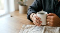 Male hands holding warm mug of coffee at wooden table with newspaper, wearing dark sweater. Morning beverage break, relaxation and leisure time at home or office workspace.