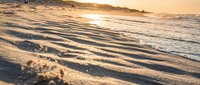 Golden sunset light illuminates rippled sand patterns on beach shore with gentle waves washing over textured surface creating peaceful coastal scene.