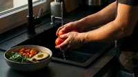 Male chef with medium skin tone washing fresh red tomato under running water at kitchen sink, preparing ingredients for cooking meal.