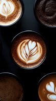 Artistically crafted latte art in multiple coffee cups showing various foam designs and patterns against dark background, highlighting barista skills and coffee culture.