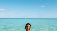 Young man with dark hair and tan skin smiling in shallow turquoise ocean water under clear blue sky, ideal for travel and vacation marketing.