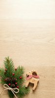 Wooden reindeer toy with checkered bow and evergreen branch decorated with red berries and pinecones on light wood surface for holiday crafts and seasonal designs.