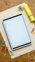 Digital tablet with blank white screen surrounded by colorful paper clips and yellow highlighter on kraft paper and wooden desk for office productivity.