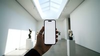 Hand holding a smartphone with blank white screen in a modern art gallery, sculptures in background, mockup ready for app or UI display.