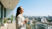 A young woman with long dark hair, appearing to be of East Asian descent, stands on a modern high-rise balcony holding a coffee cup, gazing at the cityscape below on a clear day.