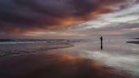 Solitary figure standing on reflective wet sand beach during dramatic sunset with purple and orange clouds reflected in water creating peaceful meditation scene.