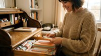 Woman with dark hair and medium complexion, wearing a beige knit sweater, organizing books in a wooden desk drawer in a home office with shelving and natural light from window.