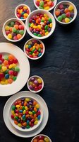 Colorful candy assortment in white bowls on dark wooden surface, featuring rainbow jellybeans, gummy bears and mixed sweets for party treats and dessert displays.