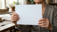 Senior man with weathered hands holding blank white paper sheet, wearing casual cardigan in home office setting with natural lighting and blurred background.