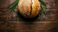 Freshly baked artisan bread loaf with decorative wheat pattern on rustic wooden table with rosemary herbs and sea salt for bakery and cooking concepts.
