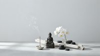 Zen spa composition with Buddha statue, white orchid, burning candle and black stones on white surface creating peaceful meditation atmosphere.