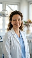 Female healthcare professional with curly brown hair and medium complexion wearing white lab coat and blue scrubs, smiling in modern medical office with bright windows and contemporary furnishings.