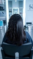 Woman with long dark hair sitting in salon chair showing hair transformation from curly to straight after professional treatment.