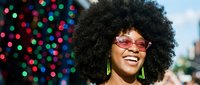 Happy African American woman with afro hairstyle and sunglasses smiling outdoors with colorful bokeh lights background for lifestyle and celebration content.