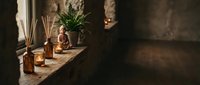 Zen meditation corner with Buddha figurine, reed diffuser, candles, and fern on rustic stone windowsill in warm ambient light.