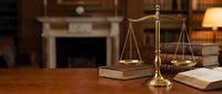 Golden scales of justice on wooden desk with law books in elegant office setting with fireplace background for legal services and attorney consultation.