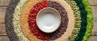 Overhead view of colorful dried legumes and grains arranged in concentric circles around empty white bowl on wooden surface for food, nutrition, and cooking concepts.