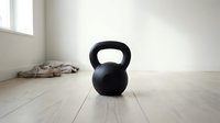 Black kettlebell weight on wooden floor in bright, minimalist gym studio space. Fitness training equipment for strength and conditioning workouts.