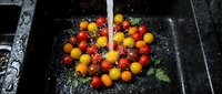 Assorted cherry tomatoes in red and yellow varieties being rinsed under running water in a dark colander, showcasing fresh produce preparation for cooking or salads.