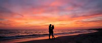 Romantic couple silhouette embracing on beach at sunset with vibrant orange and pink sky reflecting on ocean waves for wedding and love concepts.