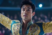 Young Asian male figure skater in ornate gold and blue sequined costume performing under spotlight, emotional expression during competition performance.