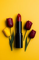 Red lipstick surrounded by burgundy and yellow tulips arranged on vibrant yellow background, creating elegant beauty and cosmetic composition for makeup artistry.