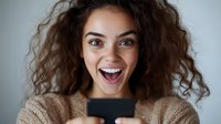 Young mixed race woman with curly hair expressing excitement while holding smartphone, showing genuine joy and surprise against light background.