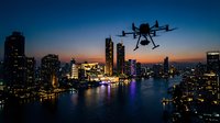 Professional drone flying over illuminated city skyline at twilight with colorful reflections on water and modern skyscrapers creating urban aerial photography scene.