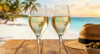 Two champagne glasses filled with sparkling wine on wooden table at tropical beach sunset with palm trees and straw hat for vacation celebration.