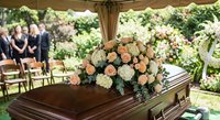Elegant funeral service with wooden casket adorned with peach and white roses under outdoor tent. Mourners gather in peaceful garden cemetery setting.