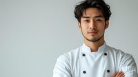 Young Asian male chef in white uniform standing with crossed arms against light background, expressing confidence and professionalism in culinary setting.