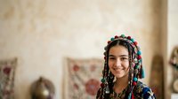 Young woman with braided hair adorned with colorful beads and ornaments, wearing traditional clothing, smiling at camera in warm interior setting.