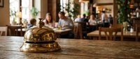 Golden brass service bell on wooden counter in busy restaurant with blurred diners and warm ambient lighting creating welcoming hospitality atmosphere.