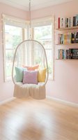 Cozy reading nook with hanging wicker chair, colorful pillows and floating bookshelves in pink bedroom corner with natural light from bay windows.