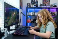 Young blonde woman gamer playing video games on computer with RGB lighting setup in modern gaming room with multiple monitors and gaming accessories.