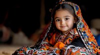 Young Asian girl wearing traditional colorful embroidered dress with orange and blue details, smiling warmly at camera against dark background.