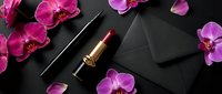 Luxury flat lay with red lipstick, black envelope, eyeliner, and pink orchid flowers on dark background, elegant beauty and fashion concept.