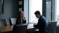 Professional business meeting with blonde woman and businessman discussing documents in modern office conference room with city view.